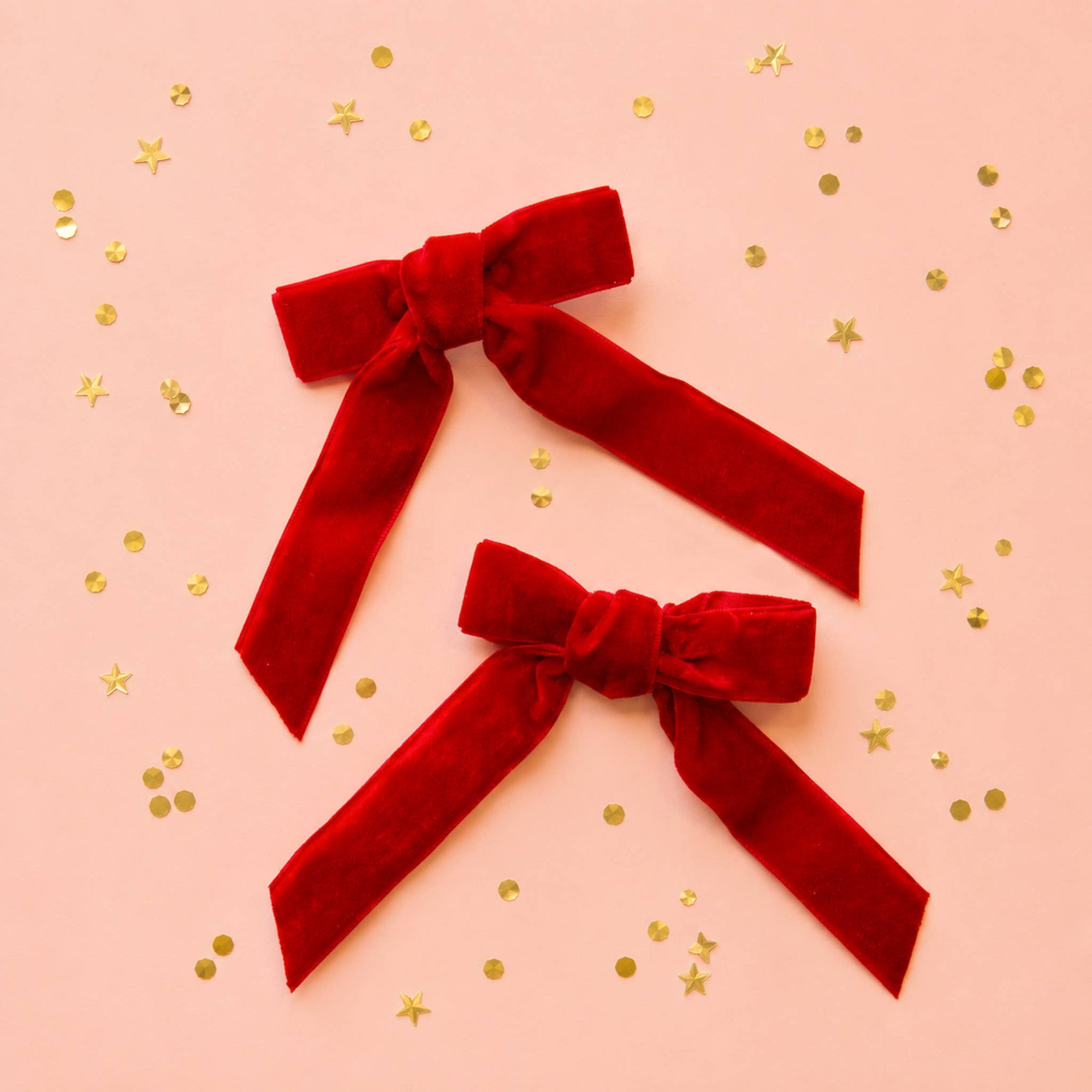 Two red velvet bow hair clips arranged on a light pink background with scattered gold confetti.