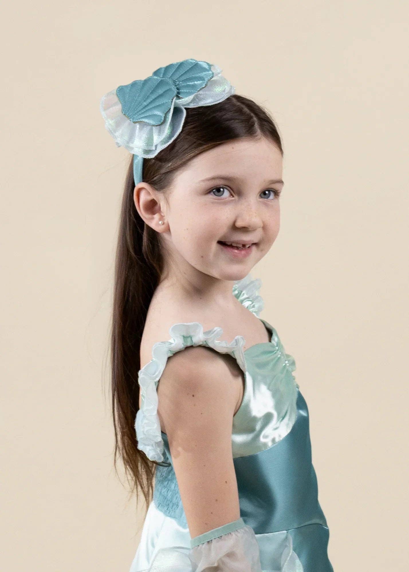 A young girl with long brown hair wearing a light blue and white ruffled costume dress and a matching light blue hair bow, photographed against a plain beige studio background.