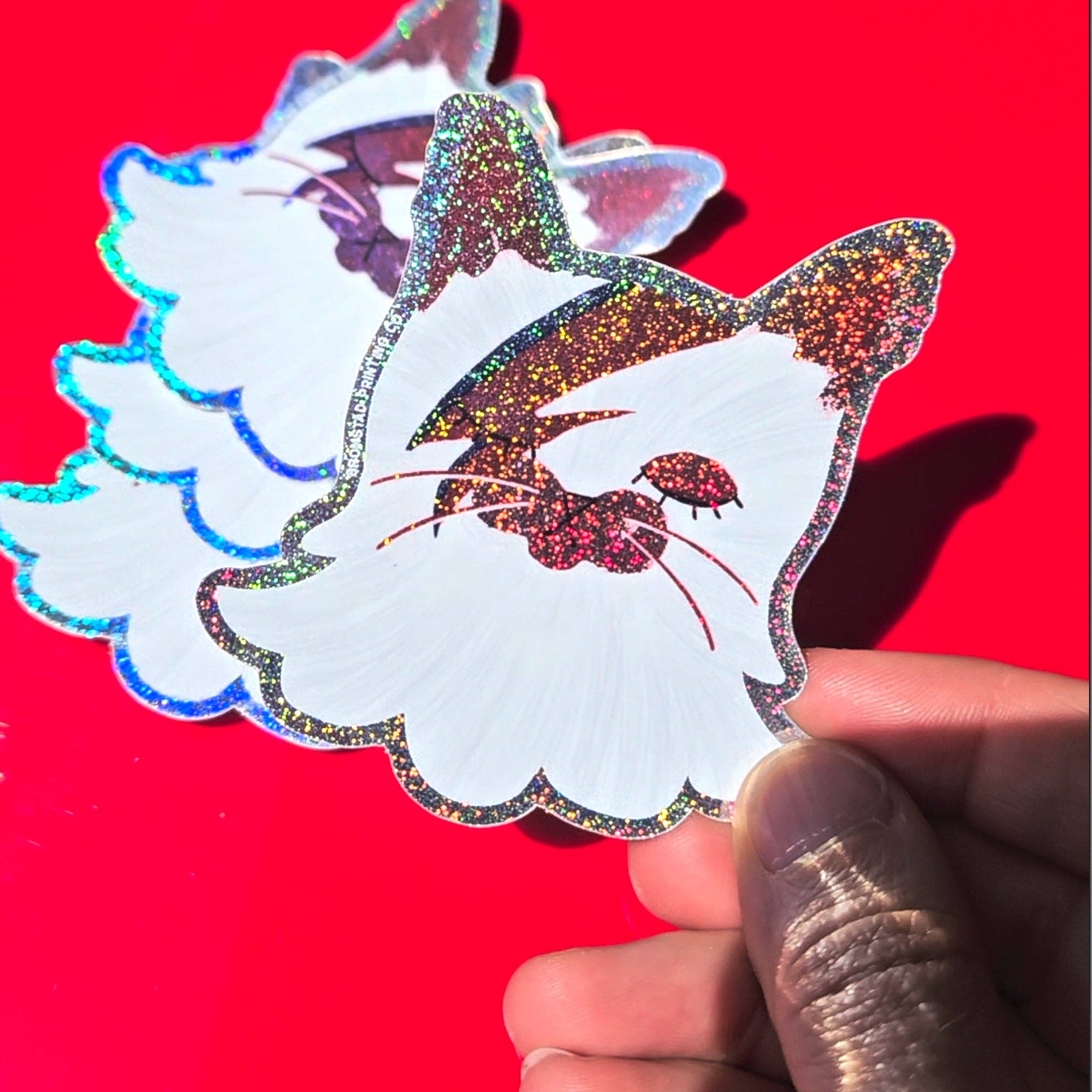 Holographic cat-shaped sticker held by a hand against a red background