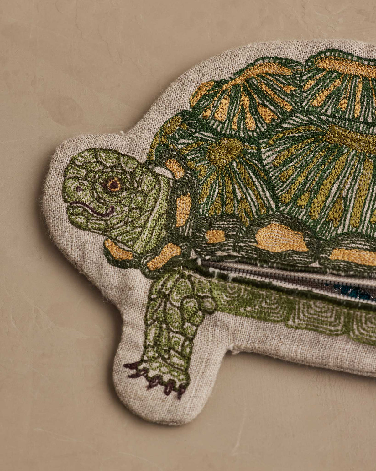 A close-up of a turtle-shaped textile patch or rug featuring an embroidered green and mustard turtle on a beige fabric base, placed on a plain beige surface.