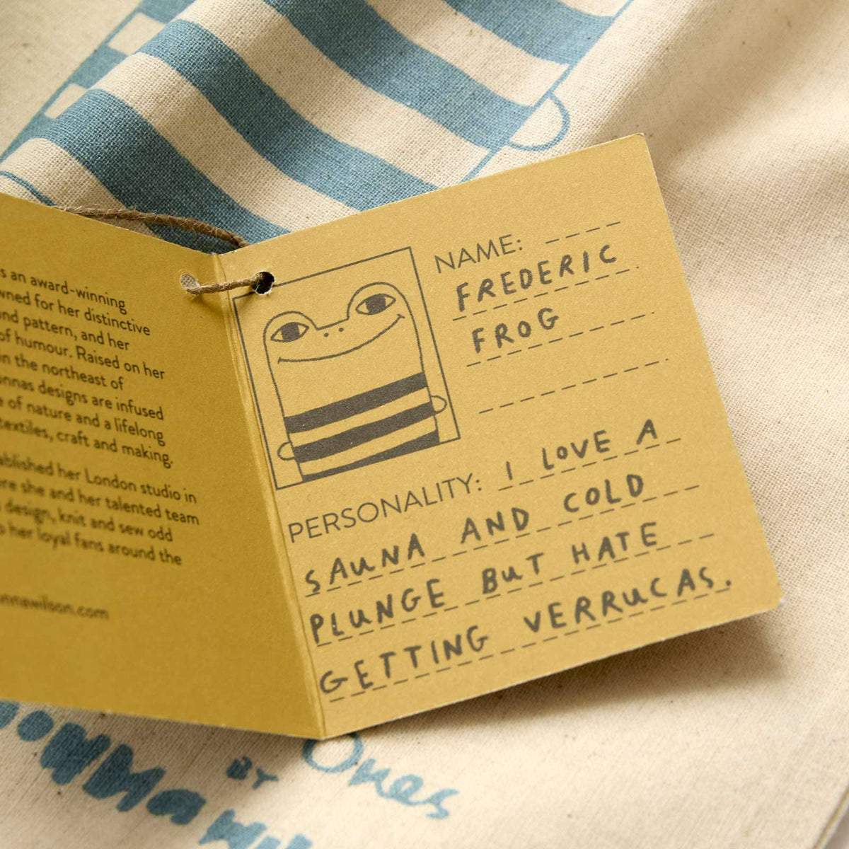 A handmade paper tag attached to a string, featuring a cartoon frog illustration and handwritten text fields including 'NAME:' and 'PERSONALITY:'. The tag is photographed on a striped fabric background.