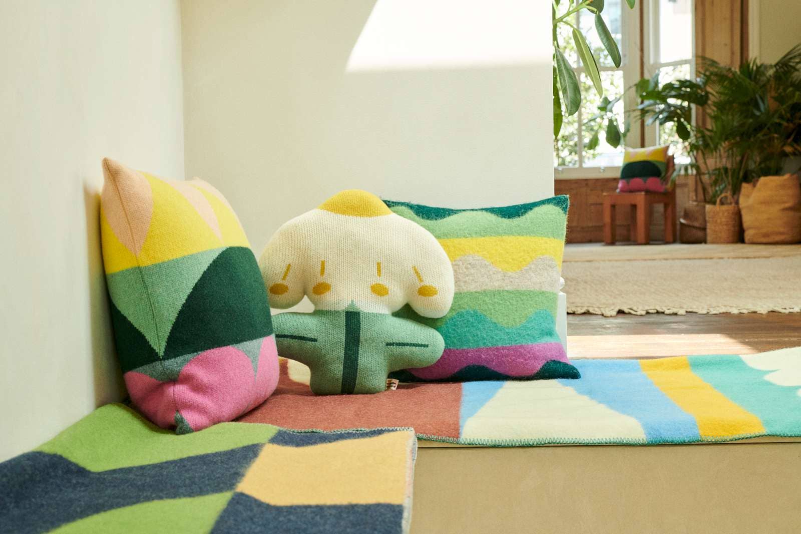 A cozy interior corner with colorful geometric throw pillows, a plush cream character pillow, and a matching multicolor rug on a wooden floor.