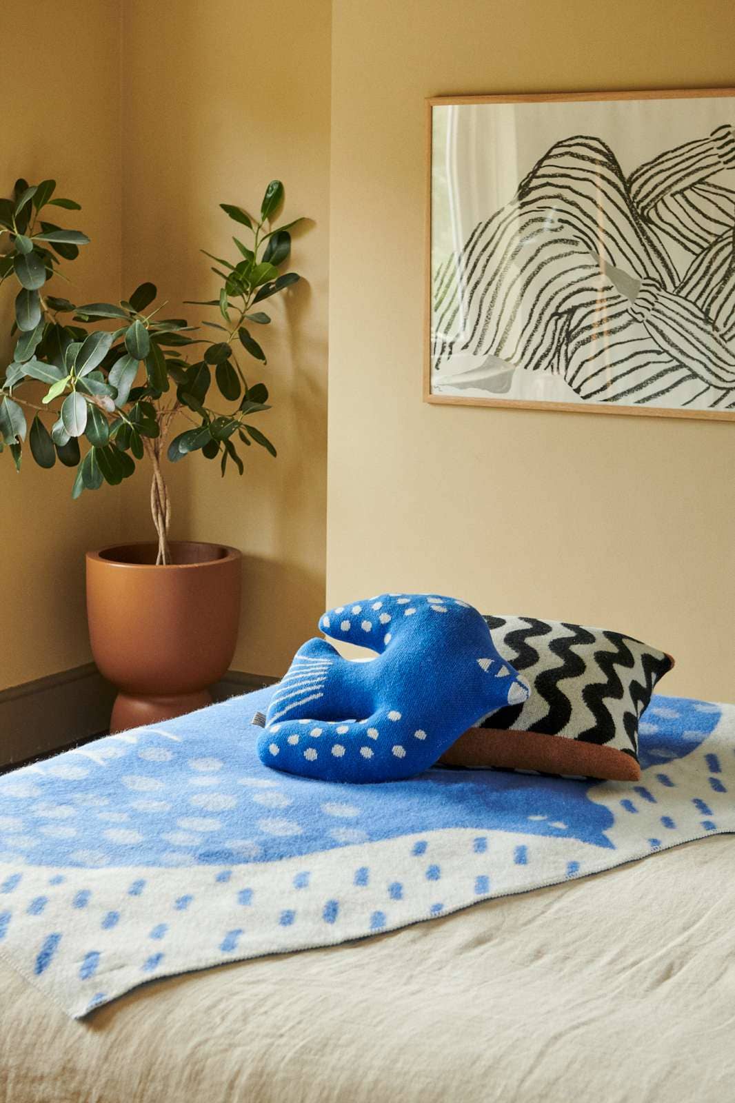 A cozy bedroom scene with a bed dressed in a blue and white patterned blanket, a bright blue textured pillow, and a black-and-white chevron accent pillow. A potted green plant stands in the corner next to a framed abstract line drawing on a warm beige wall.