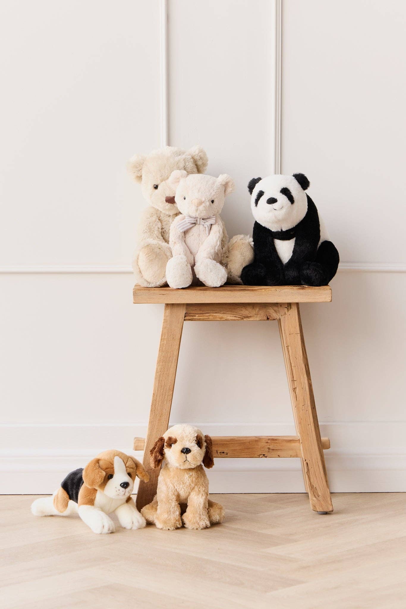 Plush toys including a panda, bear, and dog on a wooden stool against a white wall.