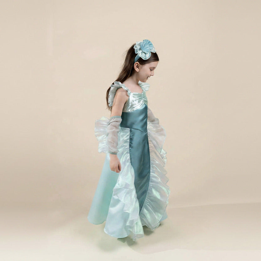 A young girl modeling a light blue and white ruffled princess costume dress with a matching blue flower headband, set against a plain beige studio background.
