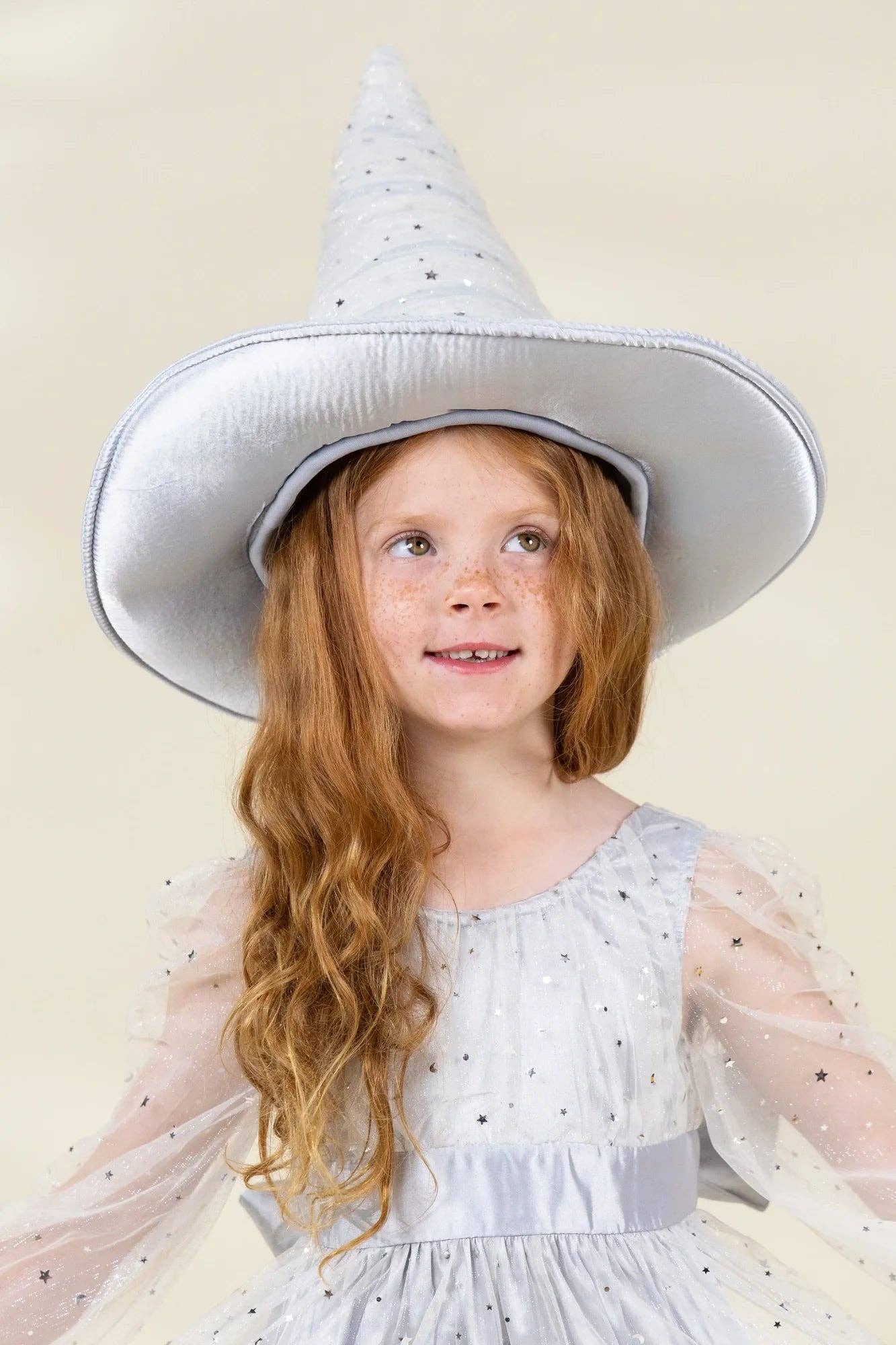 A young girl with long wavy red hair wearing a light silver-gray pointed wizard hat and a matching sheer tulle dress with small star details, set against a solid light beige background.