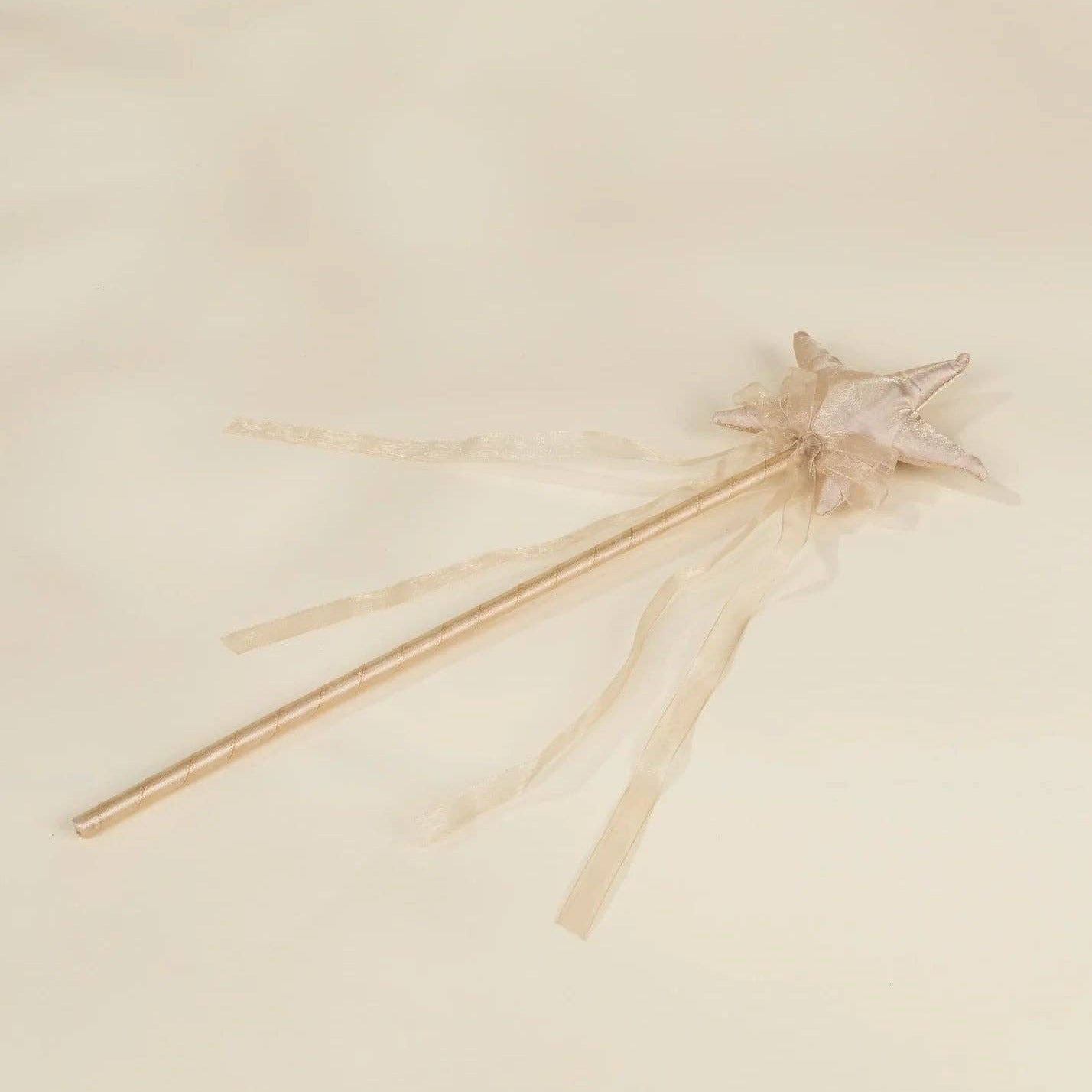 A delicate fairy wand with a gold-toned metal shaft, a small star-shaped topper, and sheer beige ribbons, resting on a soft cream-colored fabric background.