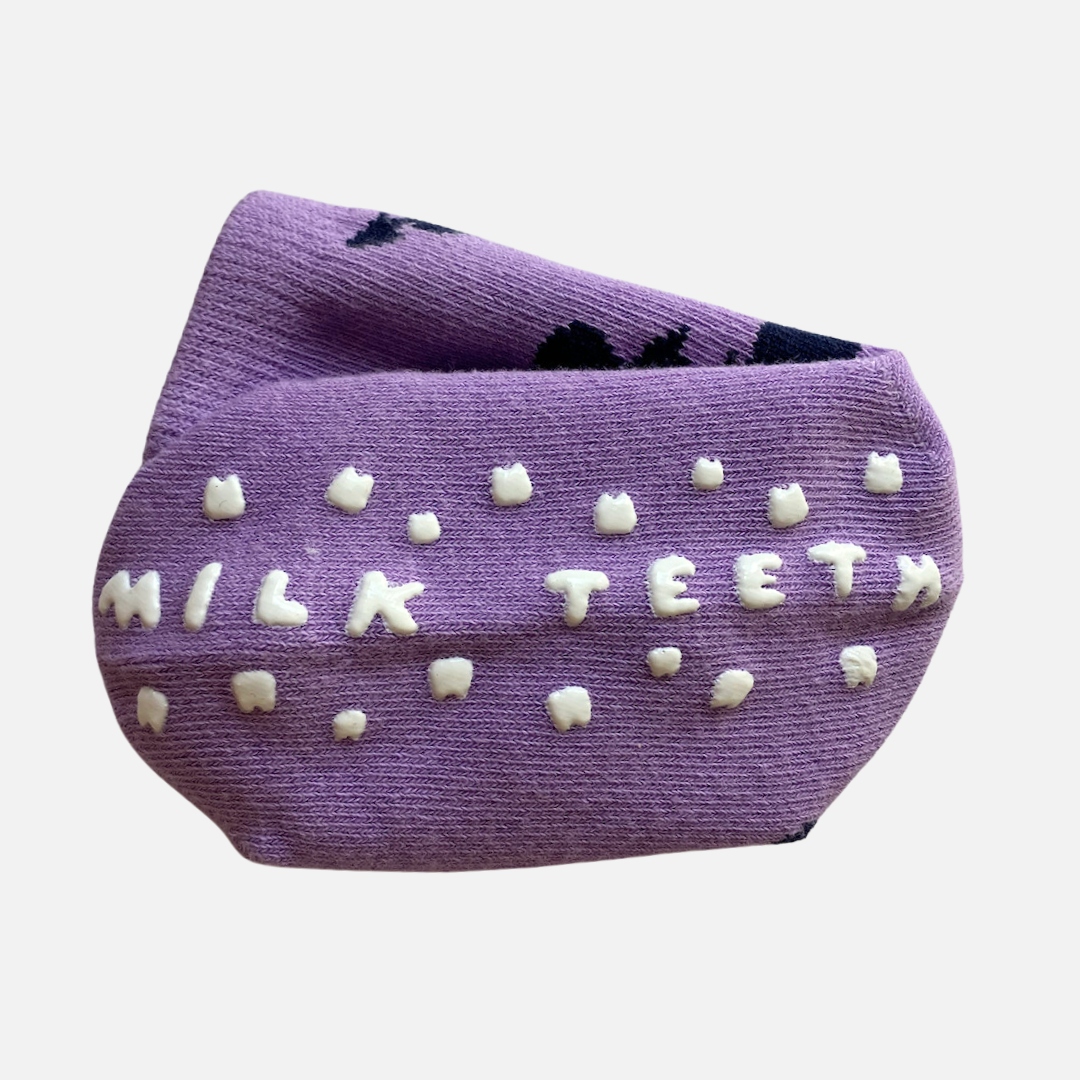 Purple socks with 'Milk Teeth' text and white polka dots on a light gray background