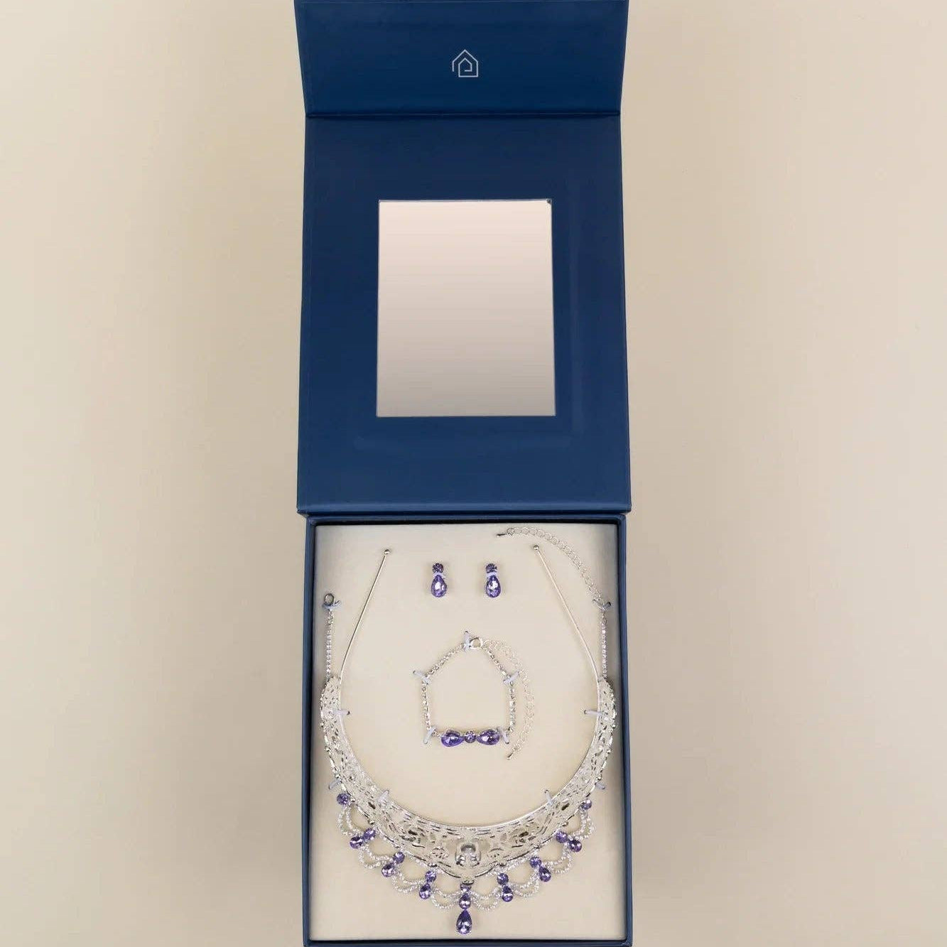 A jewelry set displayed inside an open dark blue presentation box on a light beige background. The set includes a silver-toned necklace with blue gemstone accents, matching earrings, and a small jewelry box with a mirror inside.