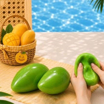 Green squishy stress toys shaped like mangoes, with two lying on a beige mat and a person's hands holding one being squeezed. In the background, a woven basket holds yellow mangoes next to a blue swimming pool.