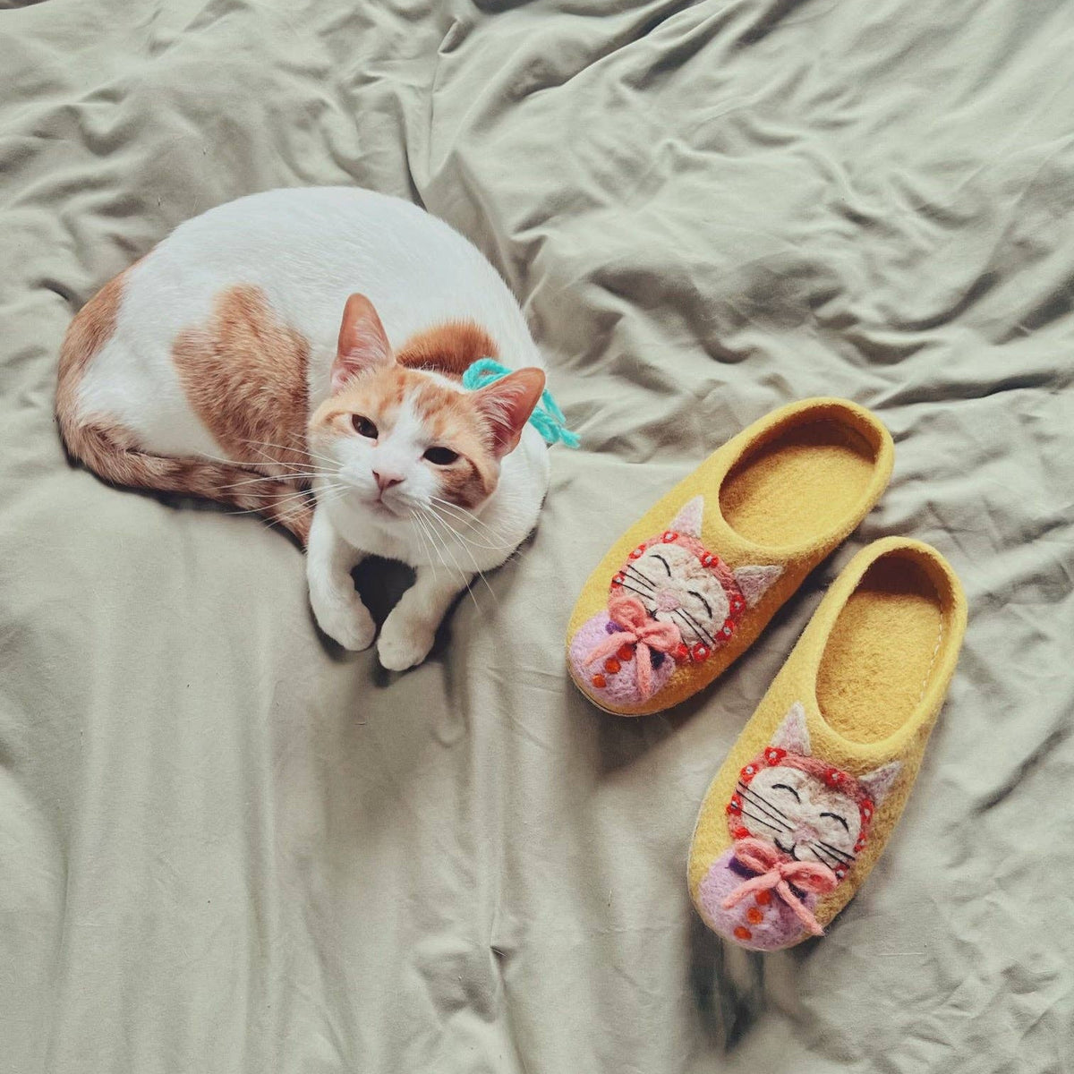 A white and orange cat lies on rumpled beige bedding next to a pair of yellow slip-on slippers decorated with a pink and gray cat face.