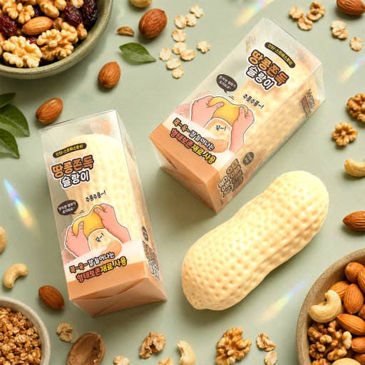 Two packaged snack products with Korean text are arranged on a pale green surface with scattered peanuts, walnuts, oats, and dried fruit pieces. The packages feature cartoon hands holding a yellow snack item, and a large peanut-shaped snack piece sits in front.