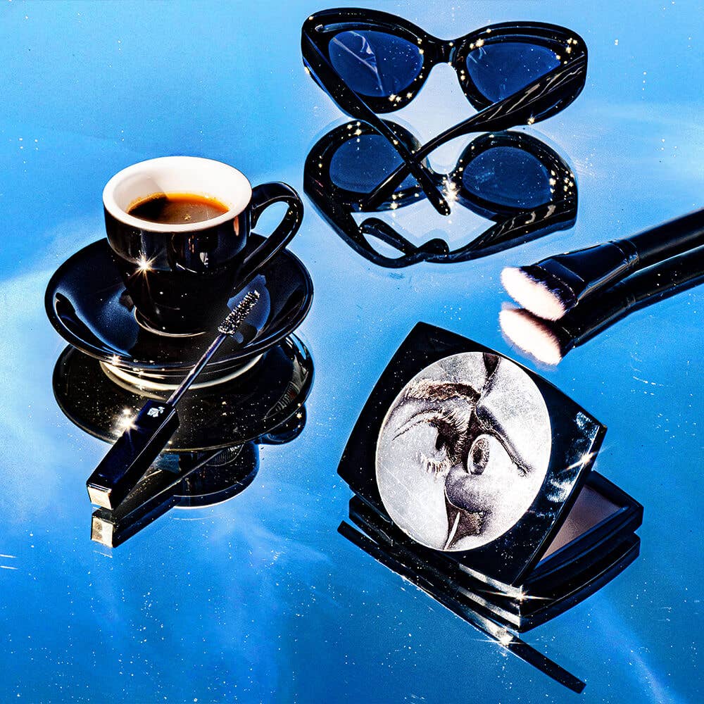 Black coffee cup, sunglasses, makeup brush, and compact mirror on a reflective blue surface