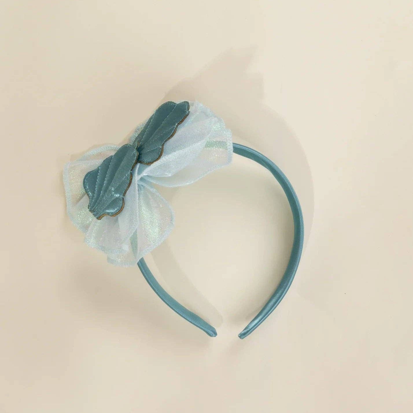 A light blue headband featuring a decorative bow made of layered tulle and a glossy, textured light blue accent piece, photographed on a solid cream background.