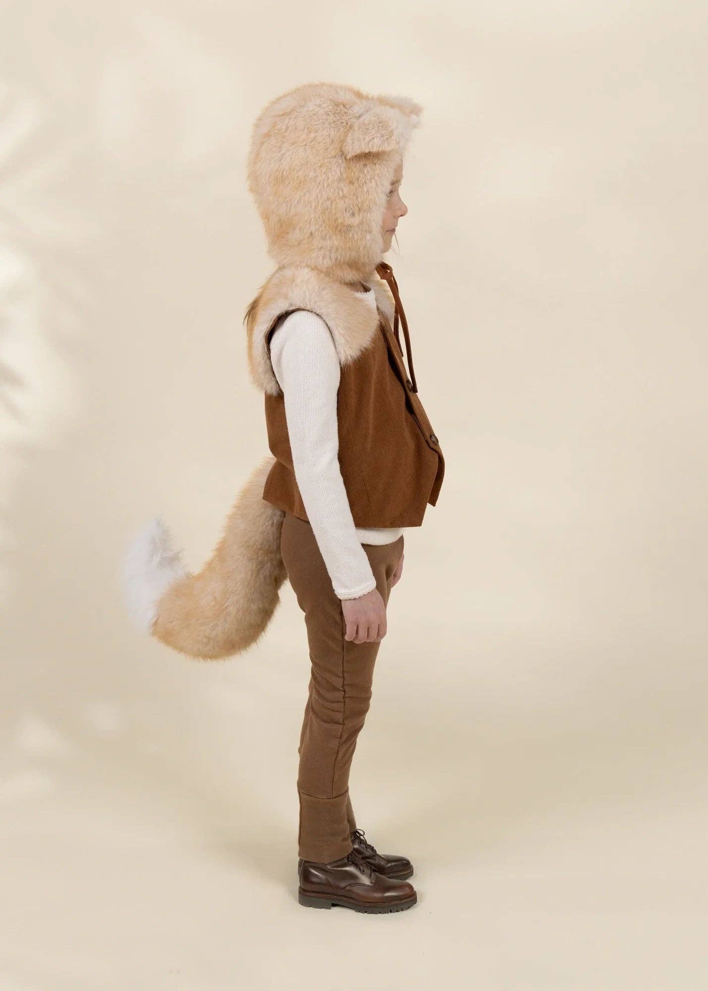 A child shown in profile wearing a brown faux-fur hooded vest with ear flaps, a long brown tail, a white long-sleeve top, brown pants, and dark brown shoes, against a plain warm beige studio background.