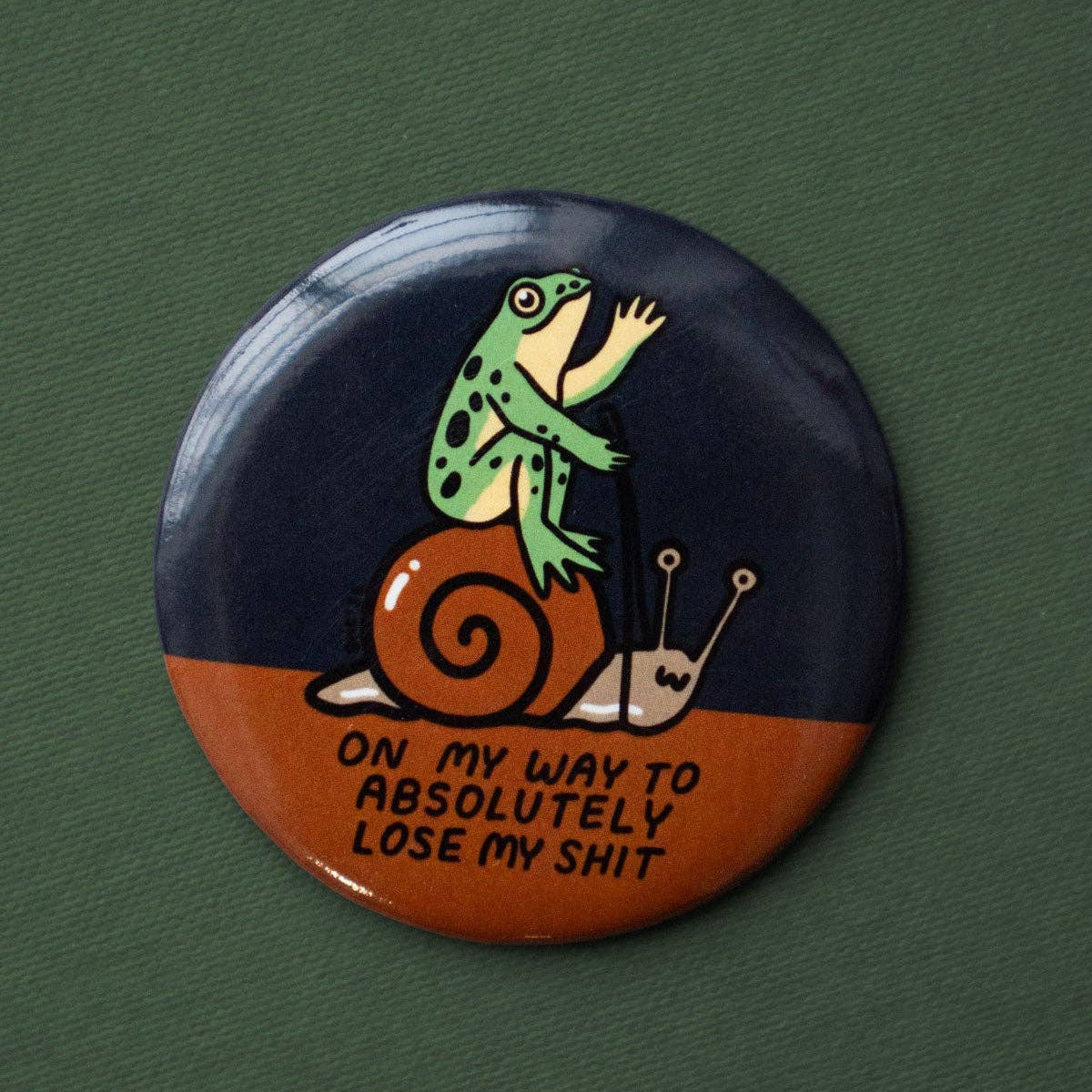 Button badge with a frog on a snail and humorous text on a green background