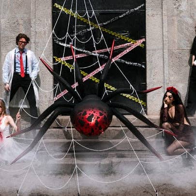 A large black spider decoration with a red and black patterned abdomen hangs in front of a stone doorway, surrounded by white fake webs, yellow caution tape, and fog. Two people in costume stand or sit nearby in a street-style setting.