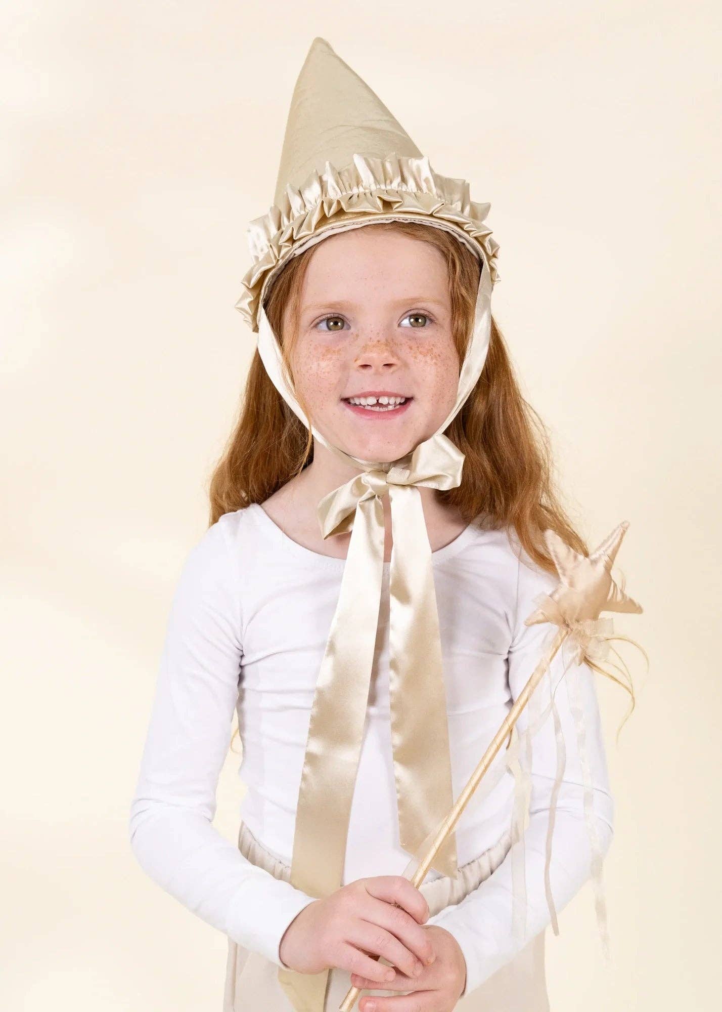 A young girl with freckles wearing a white long-sleeve dress and a beige pointed wizard hat with a large satin bow, holding a star wand against a solid beige background.
