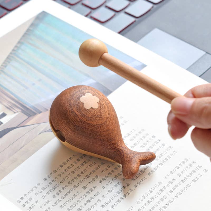 Wooden maraca being held by a hand on top of a book with a laptop in the background