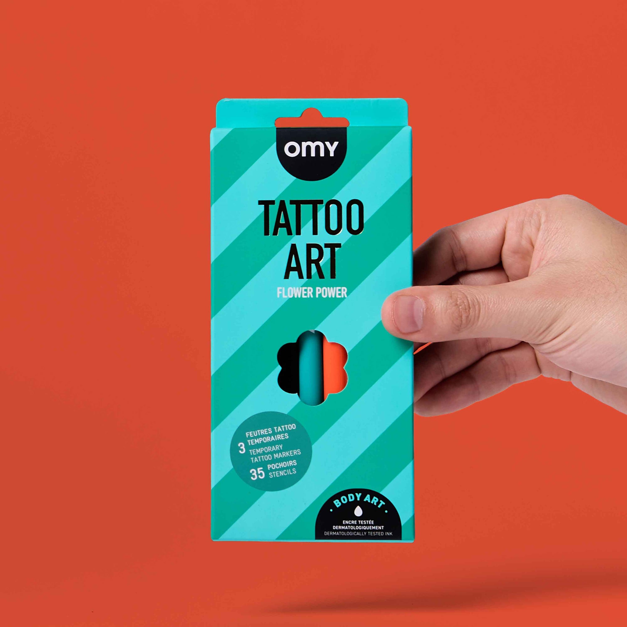 Hand holding a teal packaging of 'Tattoo Art Flower Power' by OMY against an orange background