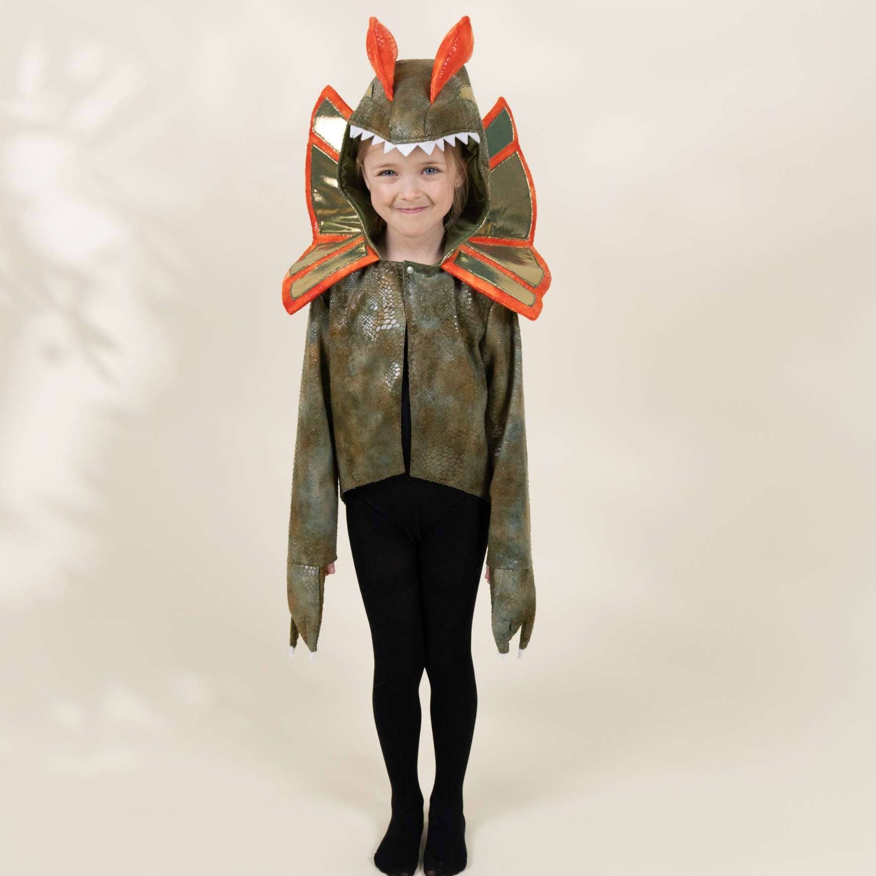 A young child modeling a dinosaur costume against a plain beige studio background. The costume includes a hood with orange dinosaur ears and teeth, a green and brown camouflage-patterned body, and orange shoulder flaps, worn over black tights.