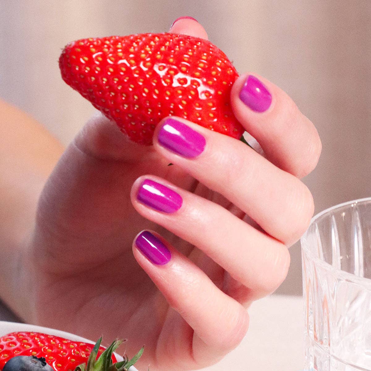 Hand with pink nail polish holding a red strawberry, blurred background