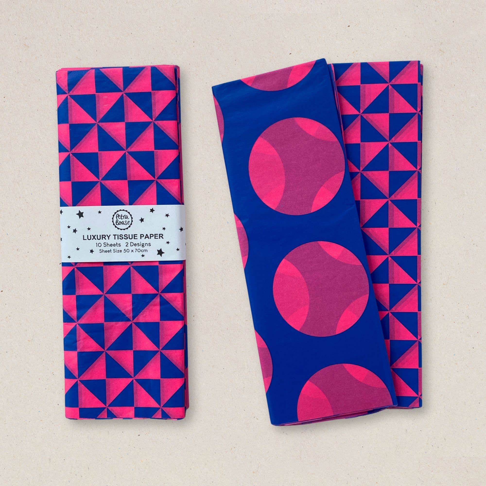 Two folded pieces of patterned tissue paper on a light beige background. One has a blue and pink geometric triangle pattern with a paper band label reading 'Luxury Tissue Paper'; the other has a blue and pink geometric triangle pattern with large pink circles.