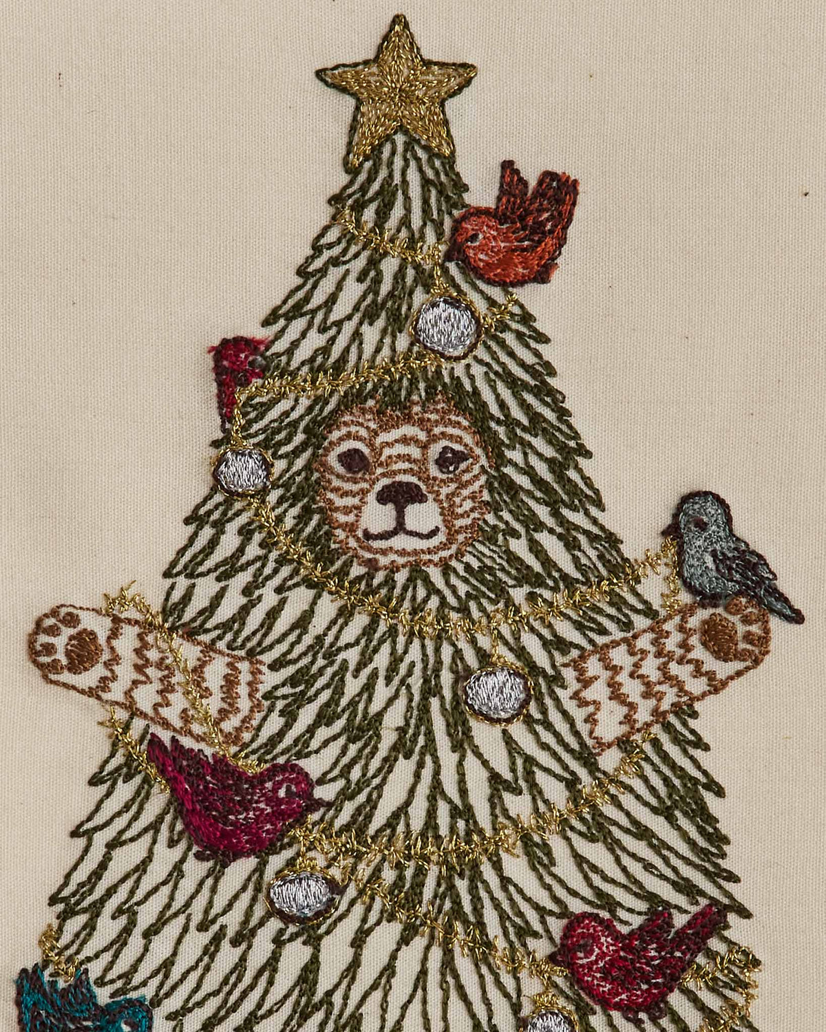 Embroidered Christmas tree design featuring a lion face in the center, decorated with gold tinsel, silver and red ornaments, birds, and a gold star on a beige fabric background.