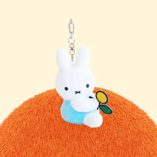 Plush keychain of a white rabbit character with a blue outfit and green leaf, sitting on an orange surface.