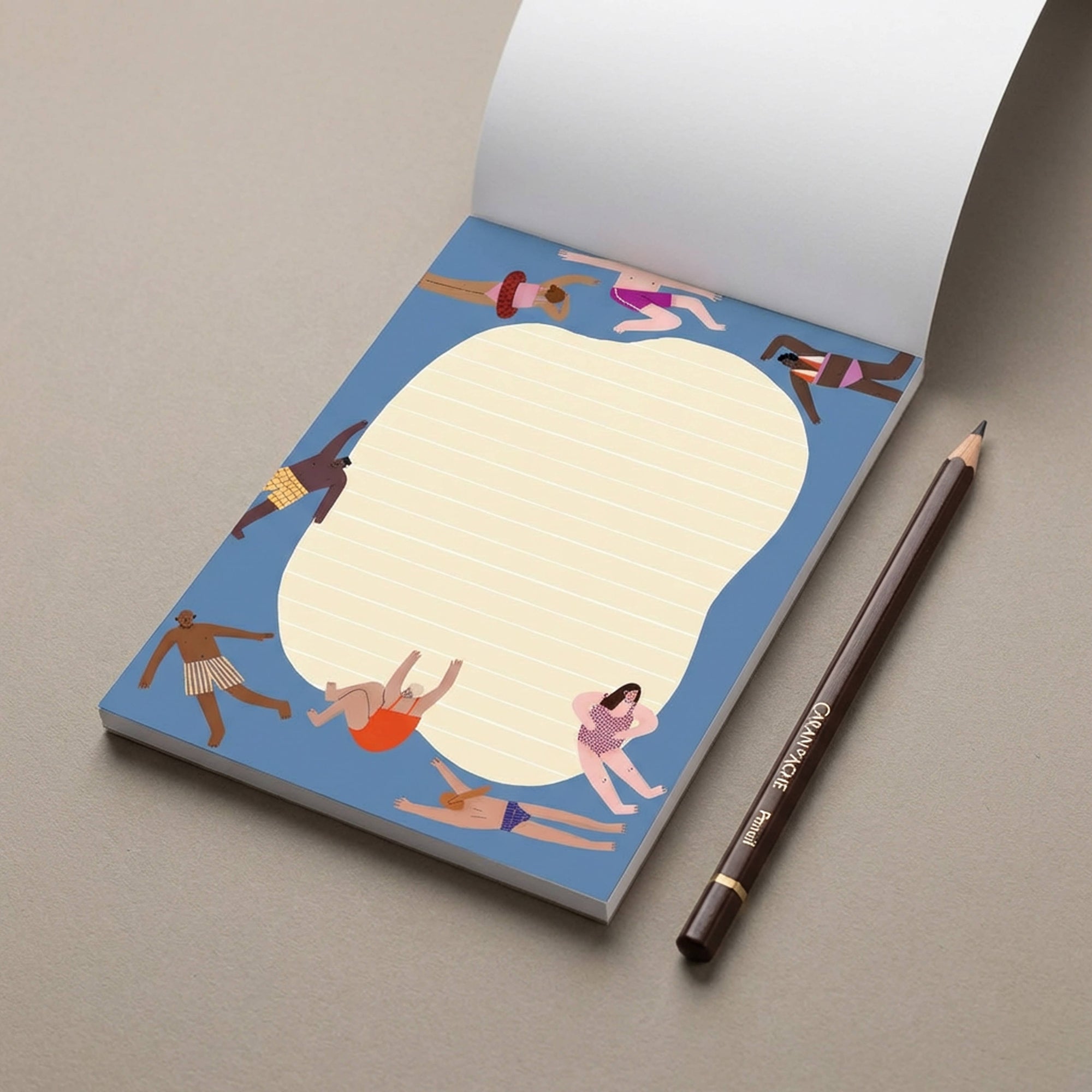 A notepad with a blue cover featuring a cartoon illustration of people sunbathing, shown open with lined paper inside, placed on a gray surface next to a brown pencil.