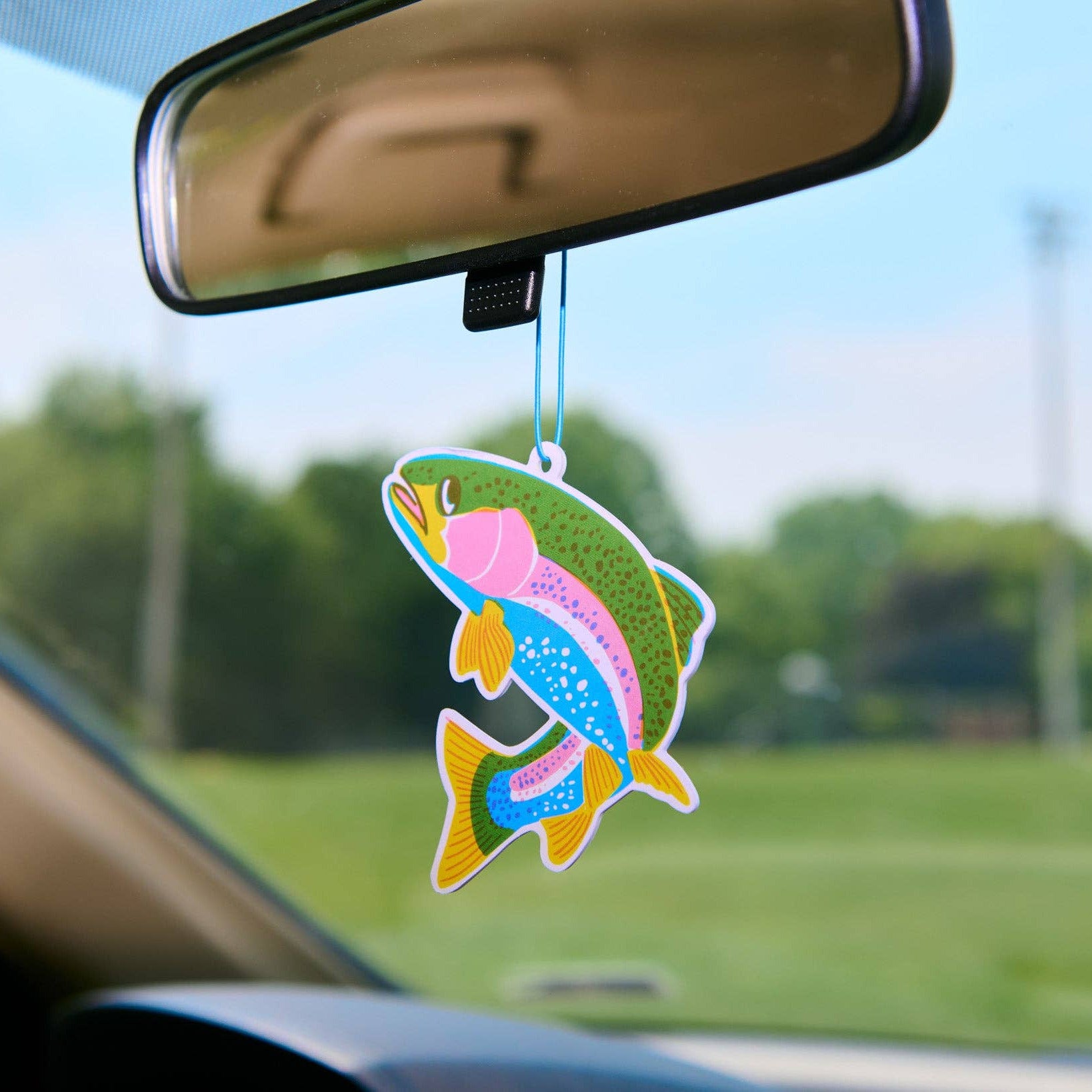 Colorful fish-shaped air freshener hanging from a car's rearview mirror with a blurred outdoor background.