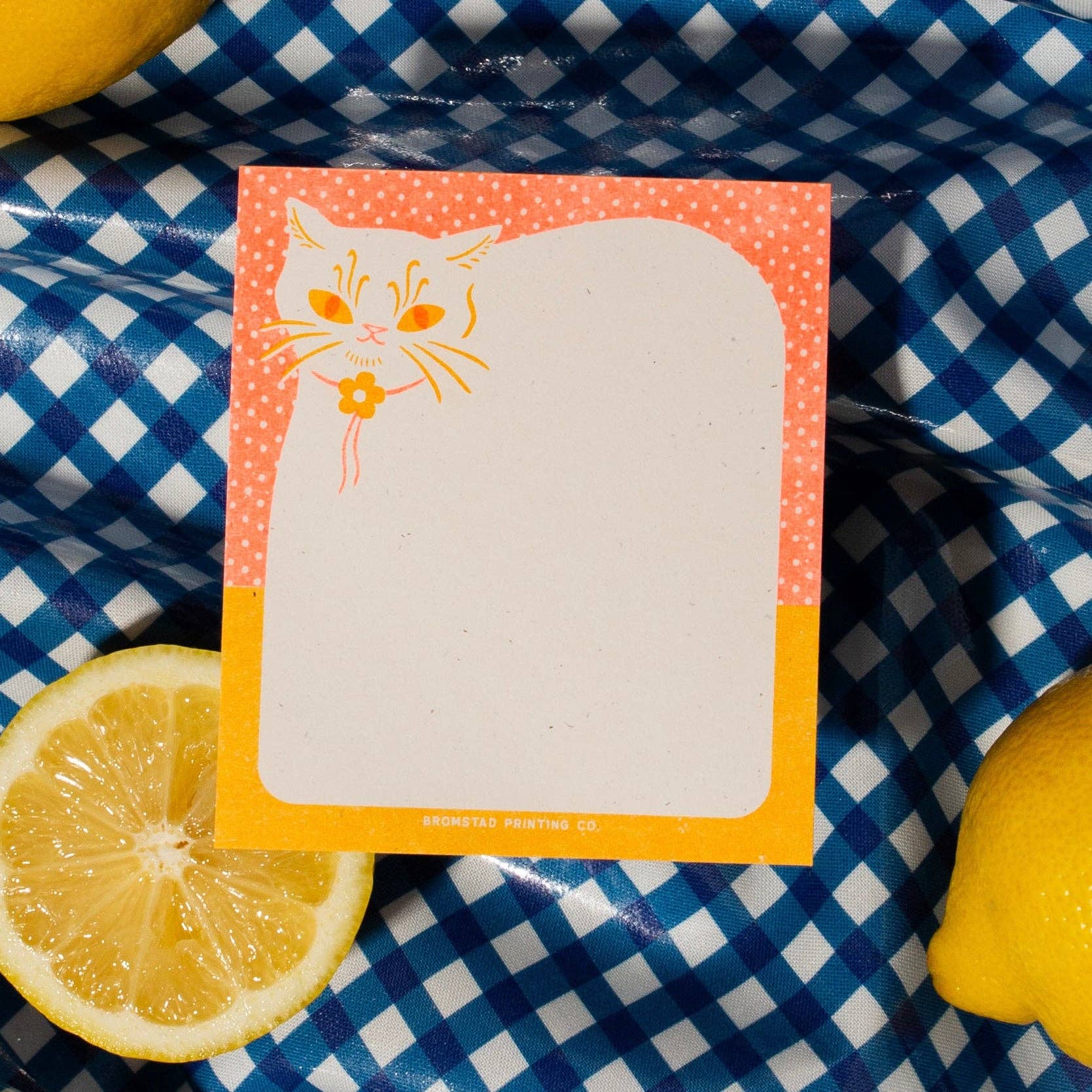 A square notepad with a white center and a coral-orange border featuring a small orange cat illustration, placed on a blue-and-white checkered fabric with lemon slices around it.
