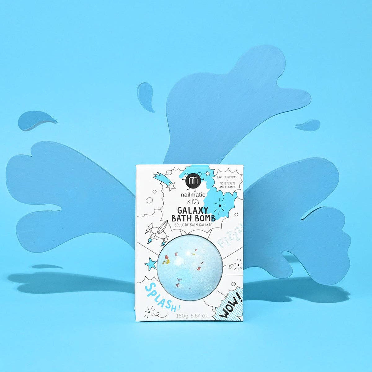 A boxed bath bomb package standing upright on a bright blue background with layered blue splash shapes behind it. The front of the box reads 'Galaxy Bath Bomb' and shows a round transparent bubble containing pastel blue and white space-themed graphics.
