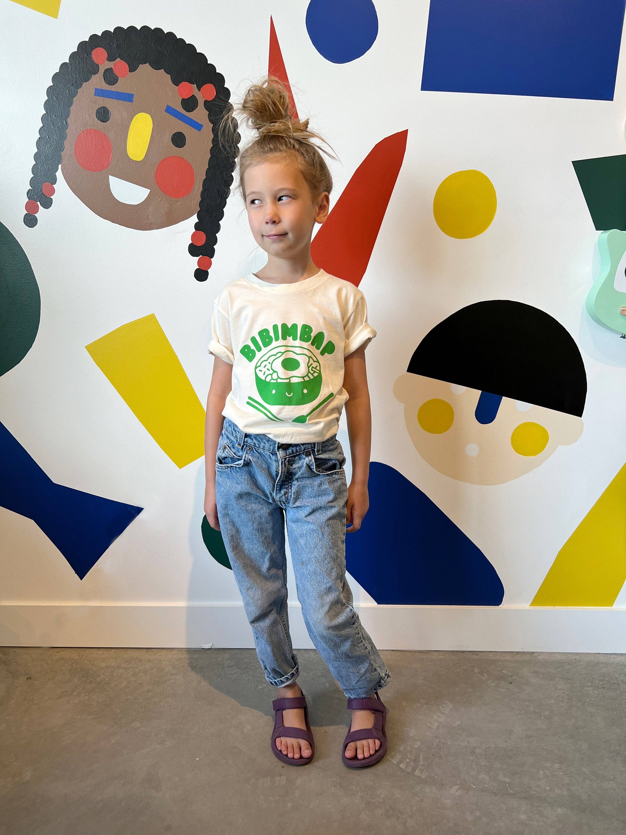Child wearing a 'Birimbar' t-shirt standing in front of a colorful abstract wall.