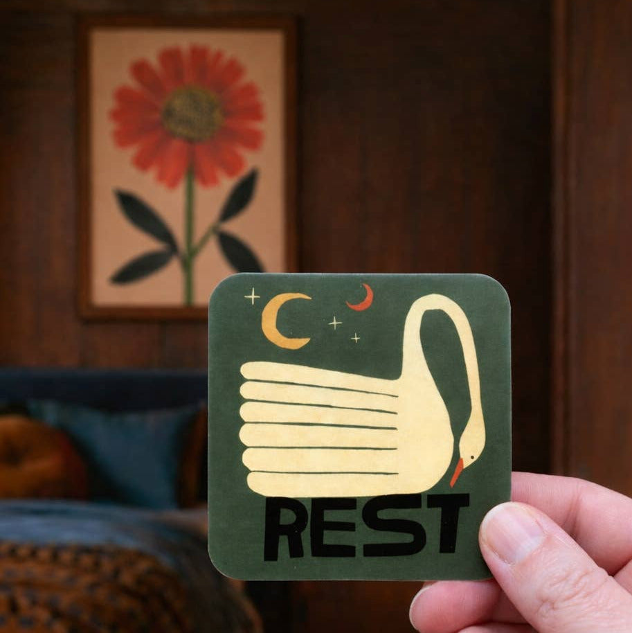 Card with a swan design and 'REST' text held by a hand, with a floral painting in the background.