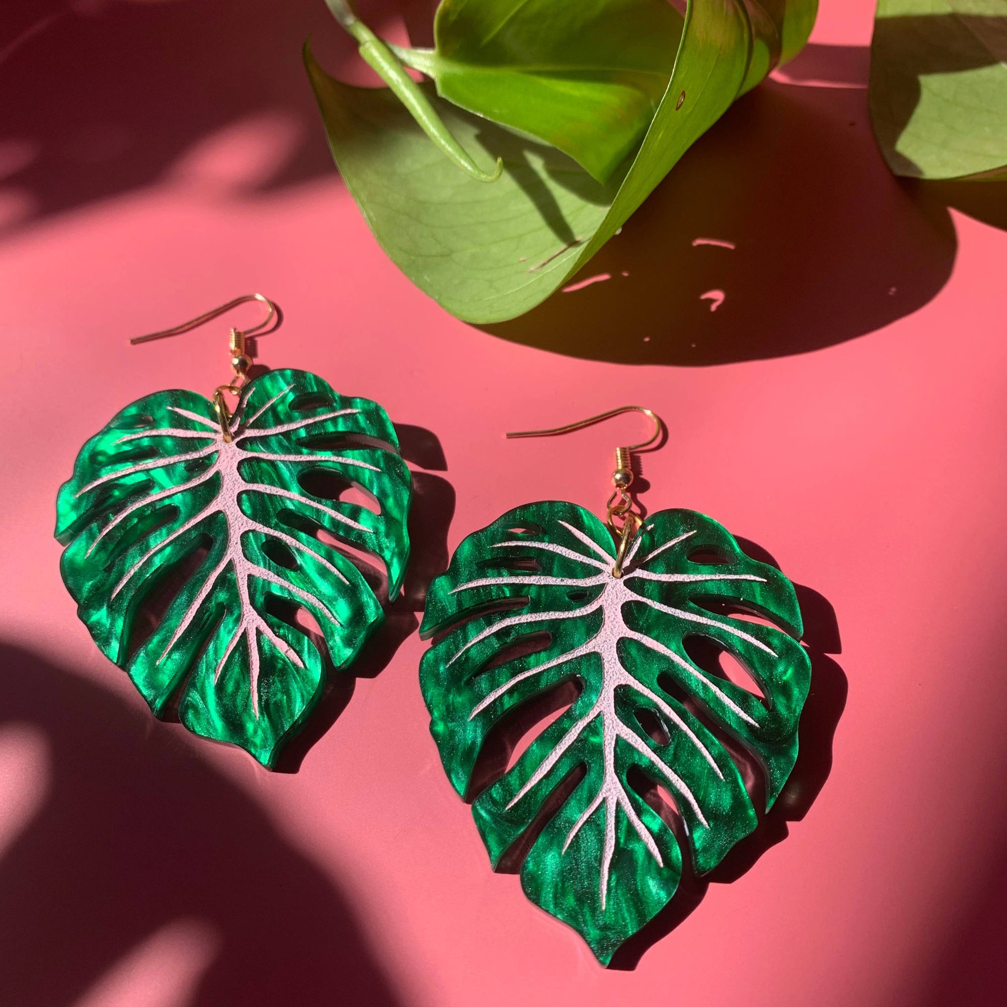 A pair of earrings designed to resemble green monstera leaves, placed on a pink surface.