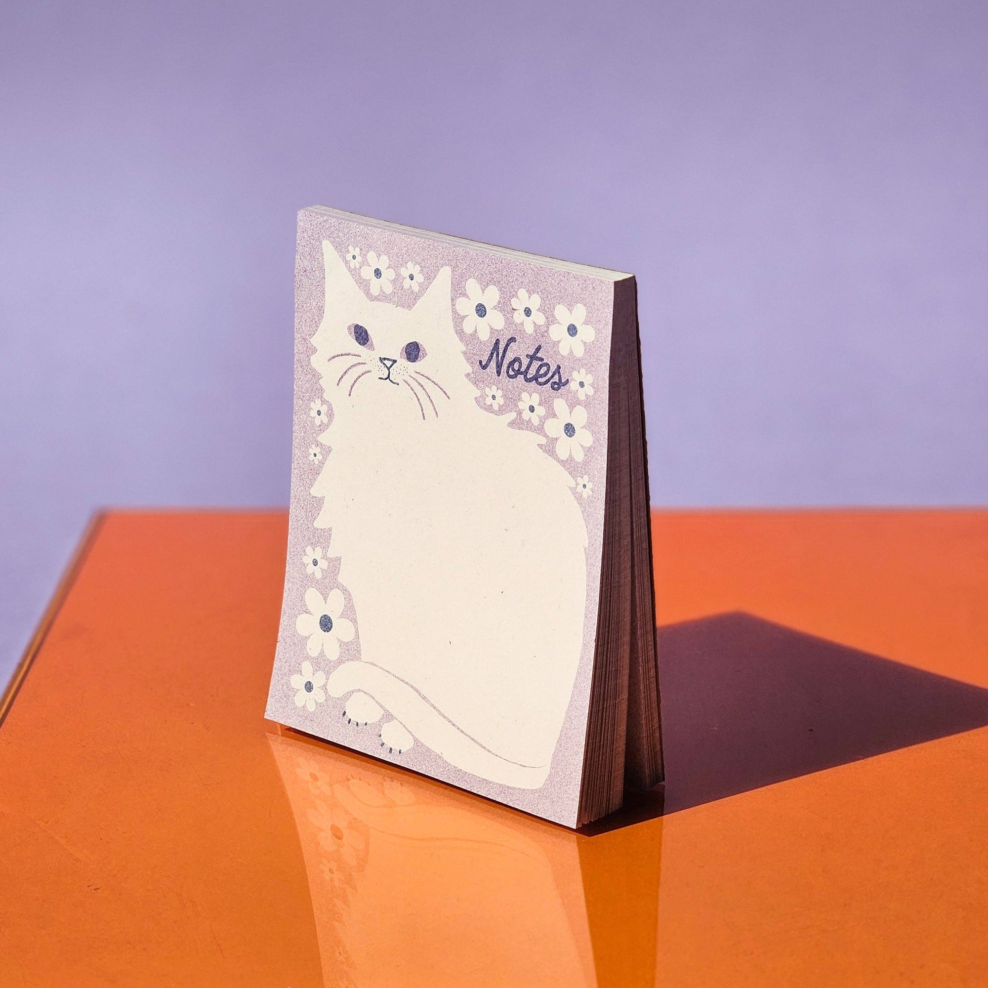 A notepad with an illustration of a cat surrounded by floral patterns, titled 'Notes', standing upright on an orange surface with a purple background.