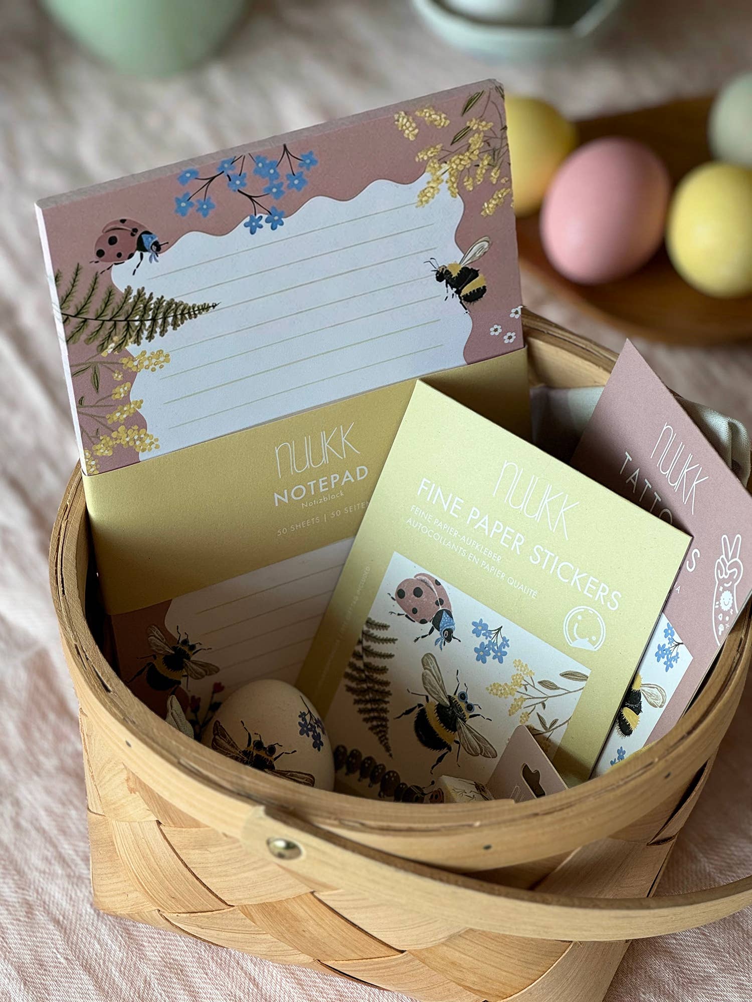 A wooden basket filled with Nukk stationery items, including a lined notepad, fine paper stickers, and sticky notes, decorated with ladybugs, bees, and ferns on a soft pink background. Colorful Easter eggs are visible in the blurred background.