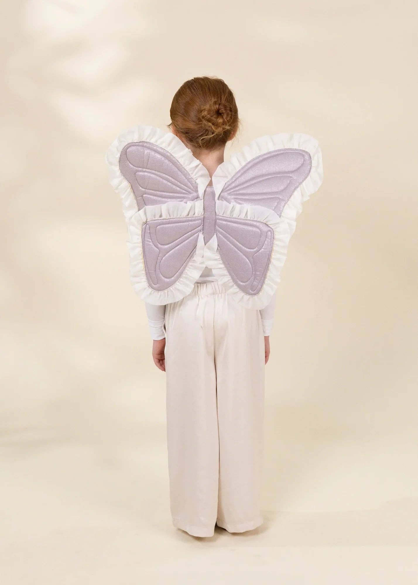 A child viewed from behind wearing a large butterfly-shaped backpiece with pale lavender quilted wings and white ruffled trim, paired with cream wide-leg pants against a soft cream studio backdrop.