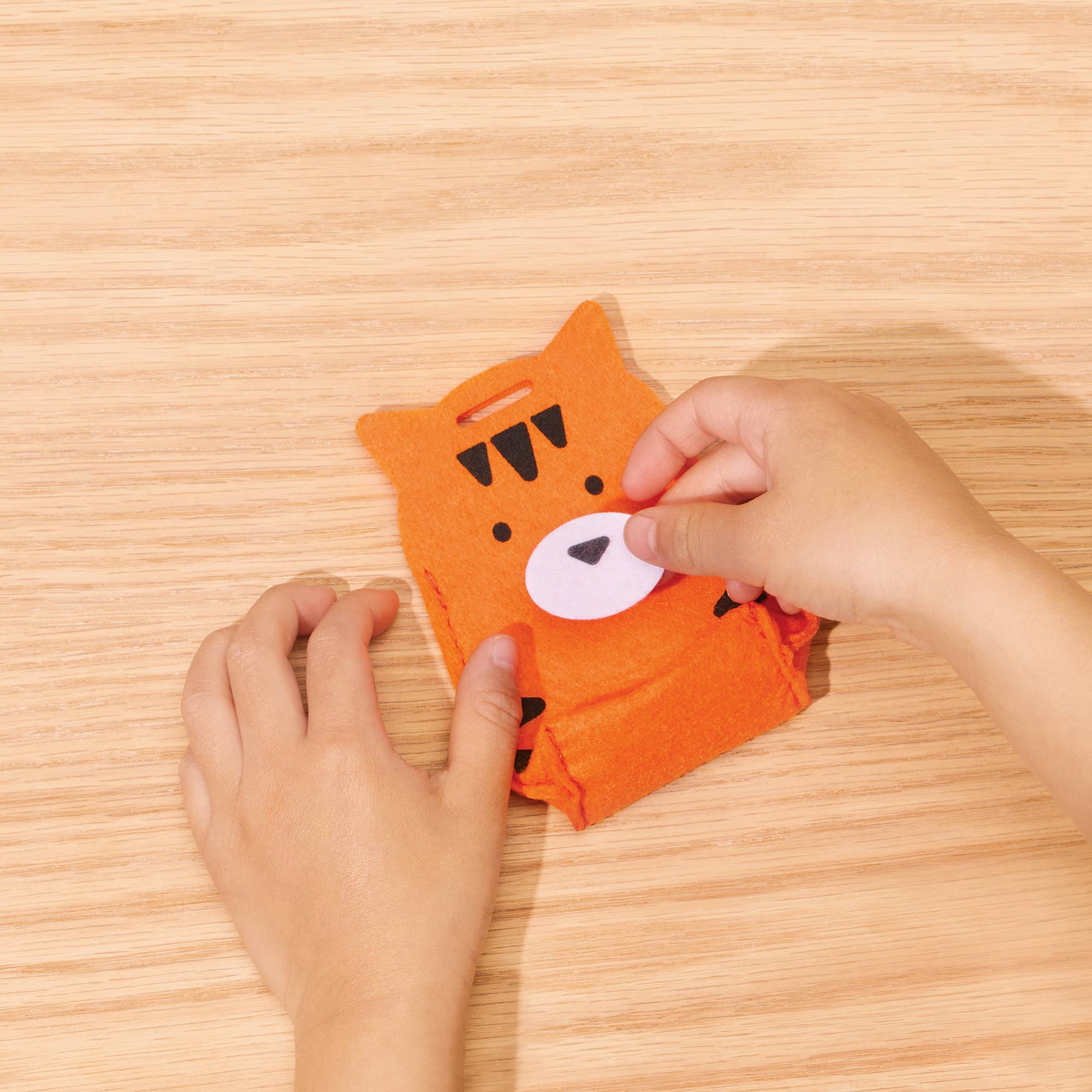 A child's hands are shown assembling a small orange felt animal pouch, likely a tiger or lion, with a white circular face patch and black embroidered details, on a light wood grain tabletop.