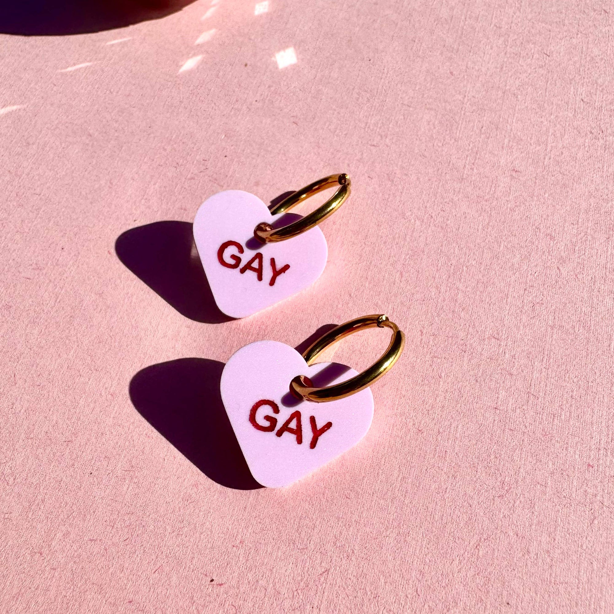 Heart-shaped earrings with 'GAY' text on a pink background