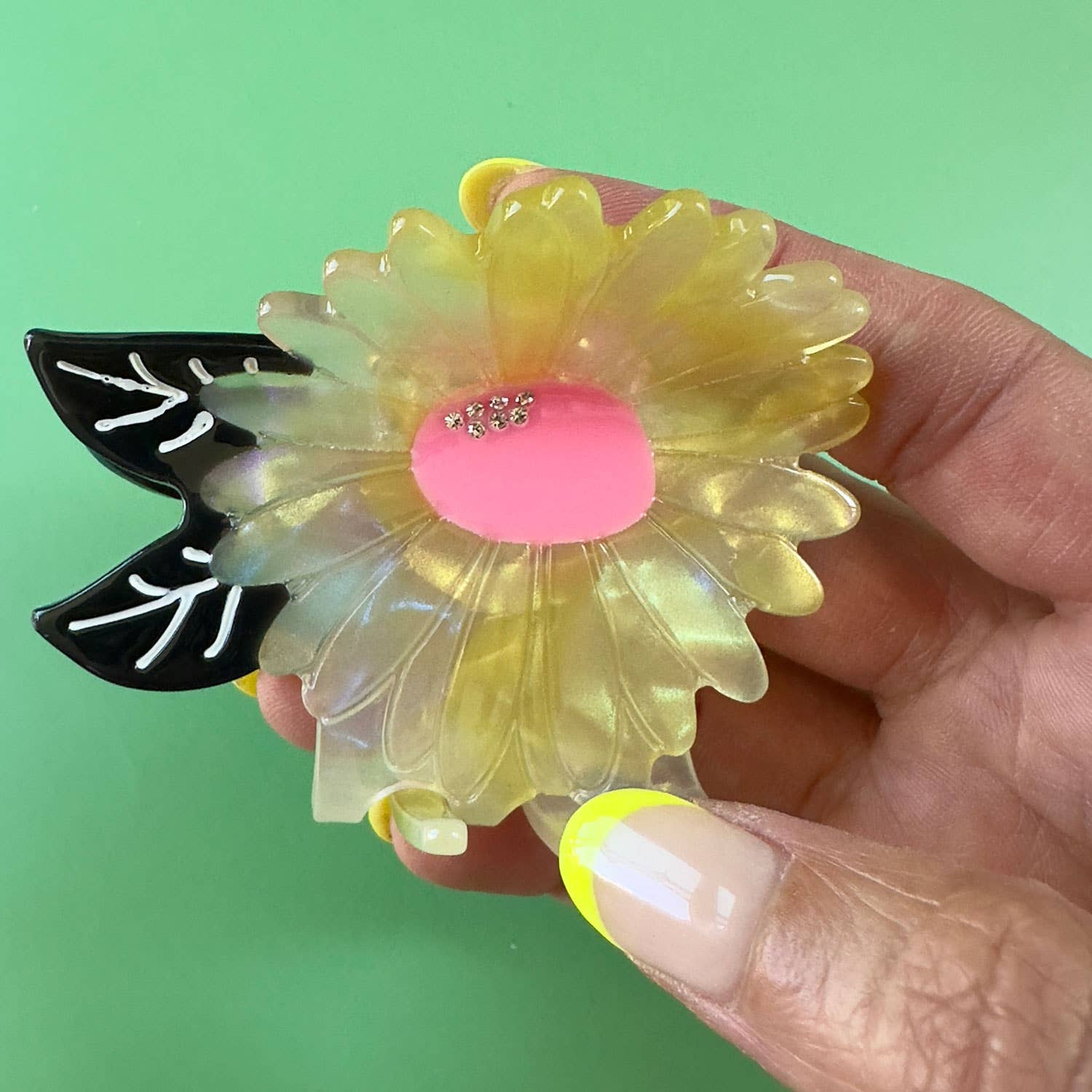 Hand holding a decorative hair clip of a yellow flower with black leaf design against a green background