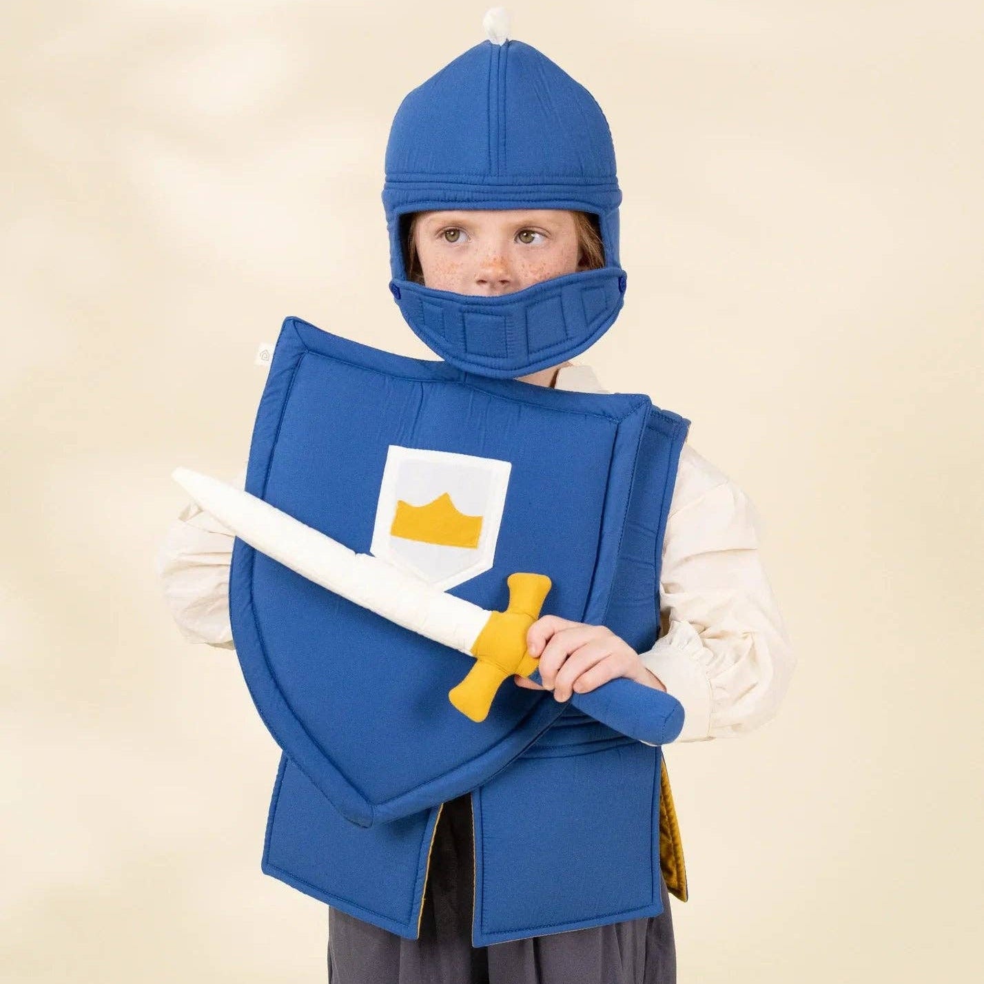 A child wearing a medieval knight costume, including a blue helmet, blue shield with a white square and yellow crown, blue vest, white tunic, and gray pants, holding a toy sword against a warm beige studio background.