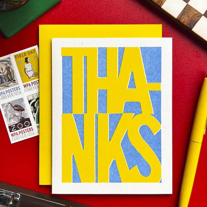 A typographic art print card reading 'THANKS' in large yellow letters over a blue textured background, placed on top of a yellow envelope on a red surface with postcards and a yellow pen nearby.
