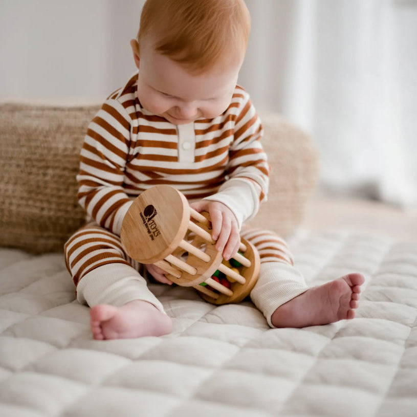 A baby sitting on a quilted cream mattress, wearing a brown and white striped long-sleeve romper and holding a round wooden baby toy 