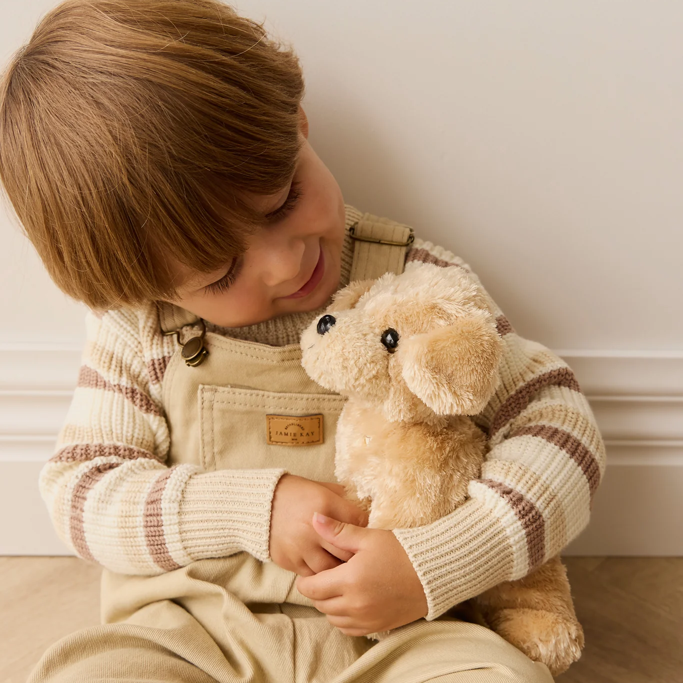 Child wearing a beige outfit holding a teddy bear in a cozy indoor setting