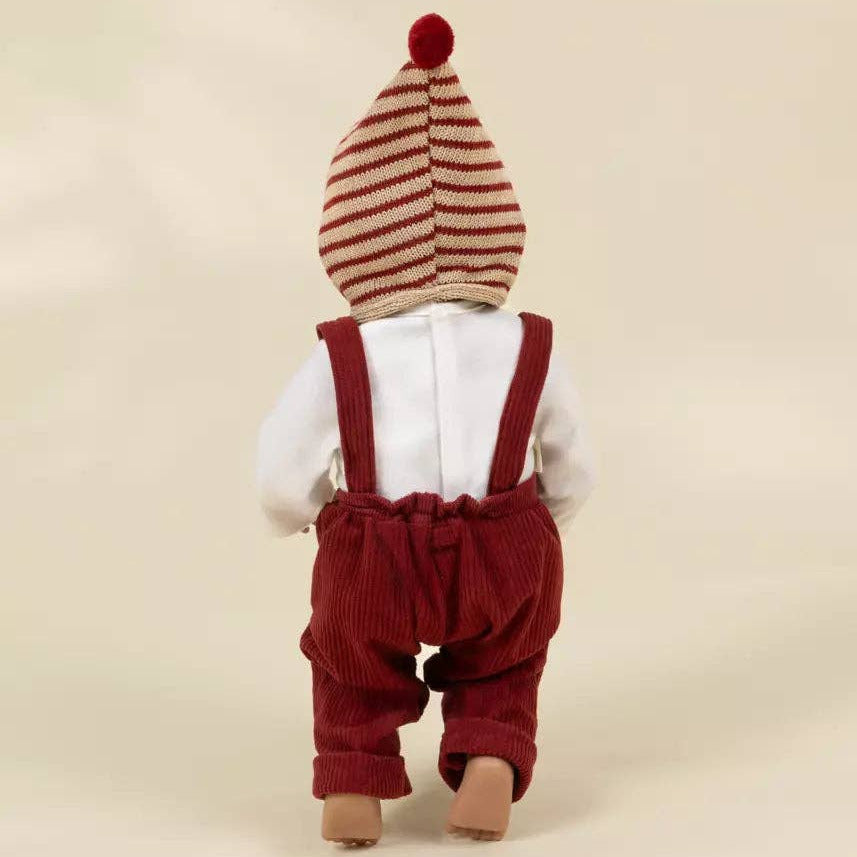 A baby doll viewed from the back, wearing a cream and red striped knit hat with a red pom-pom, a white long-sleeve shirt, and red corduroy suspenders and pants, set against a plain beige background.