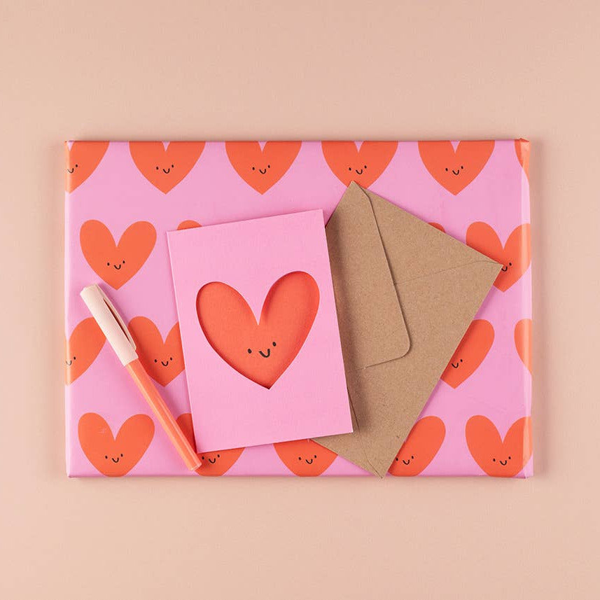 Card with a heart design on a pink background with more hearts, a pen, and envelope.