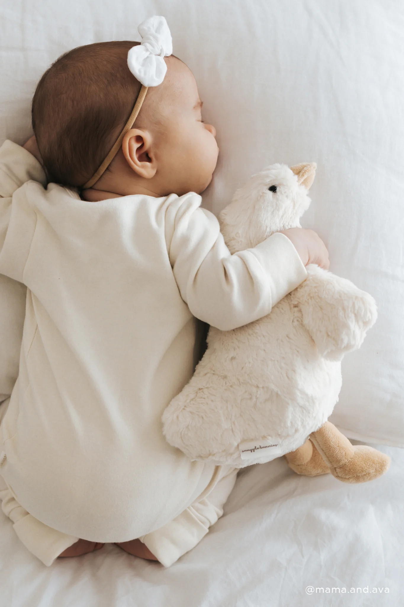 Baby in a white outfit holding a plush toy, wearing a headband with a bow, on a white background.