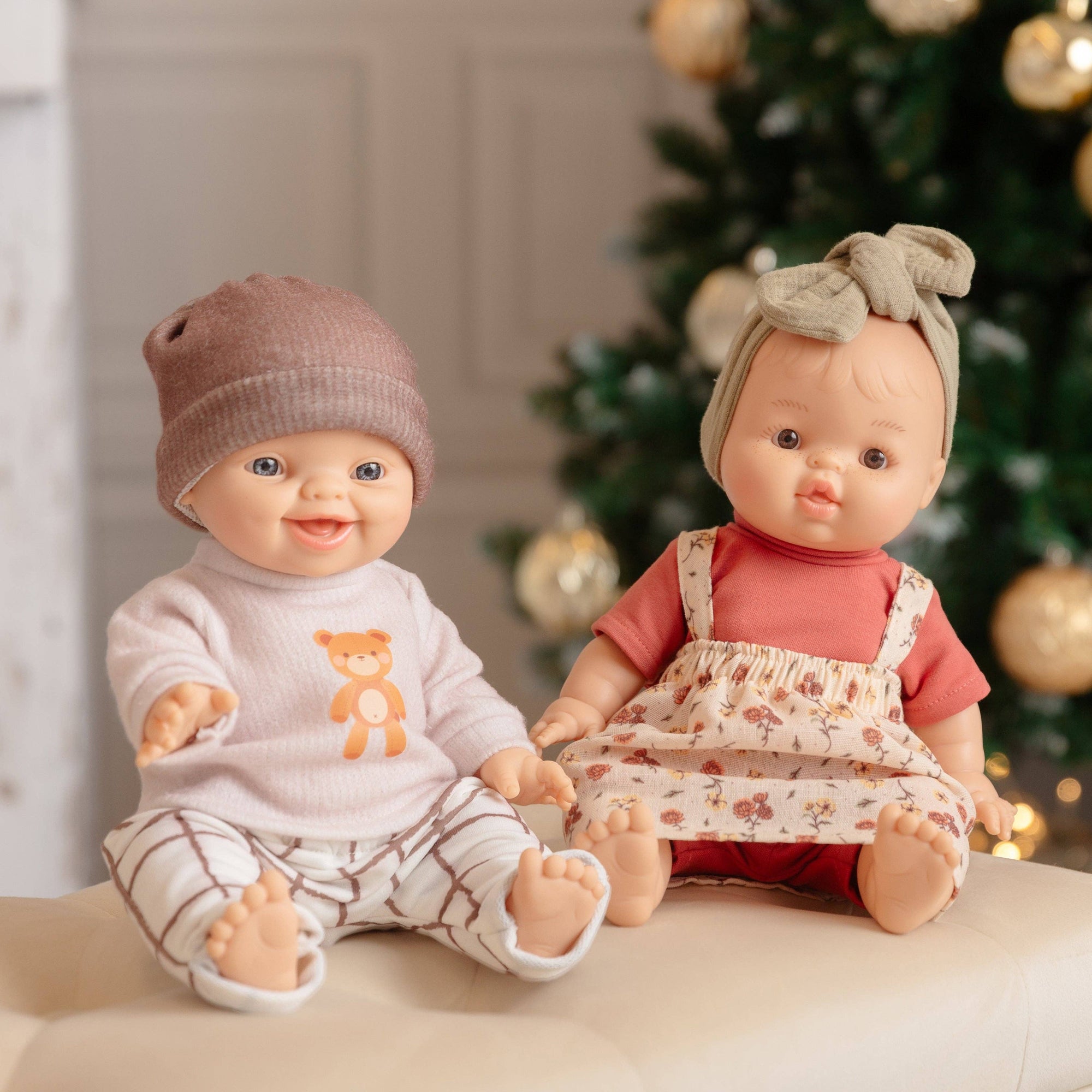 Two dolls sitting on a surface with a decorated Christmas tree in the background.